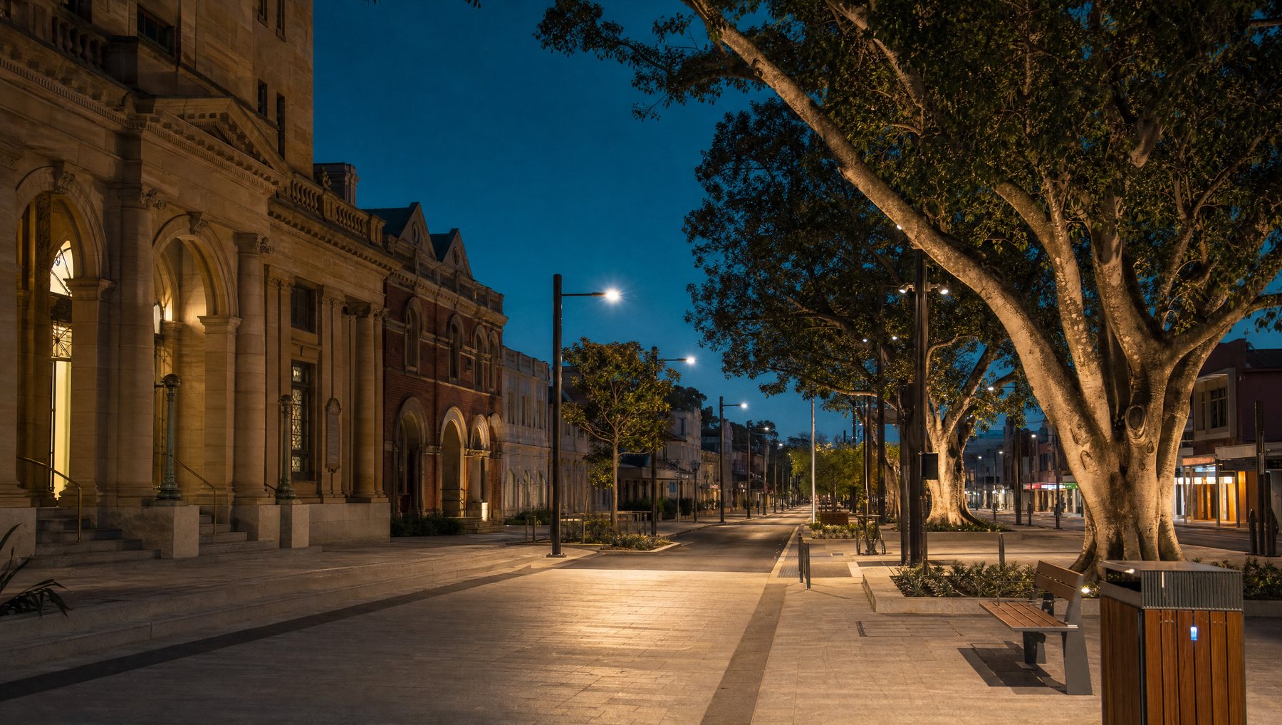 A civic streetscape at dusk with heritage buildings and street lighting switched on