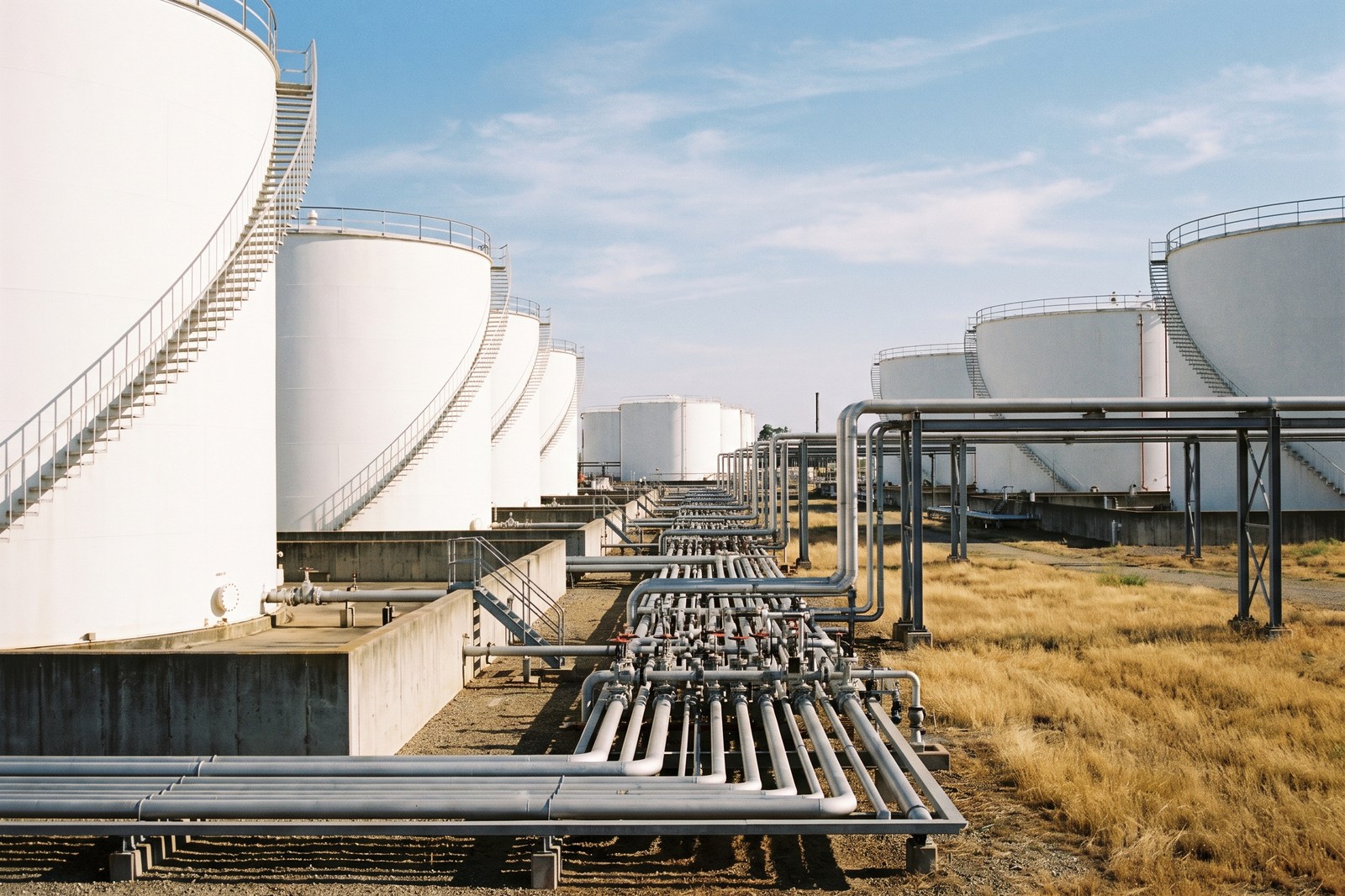 A refinery fuel storage tank farm, white cylindrical above-ground tanks with external spiral stairways and pipework manifolds on steel supports