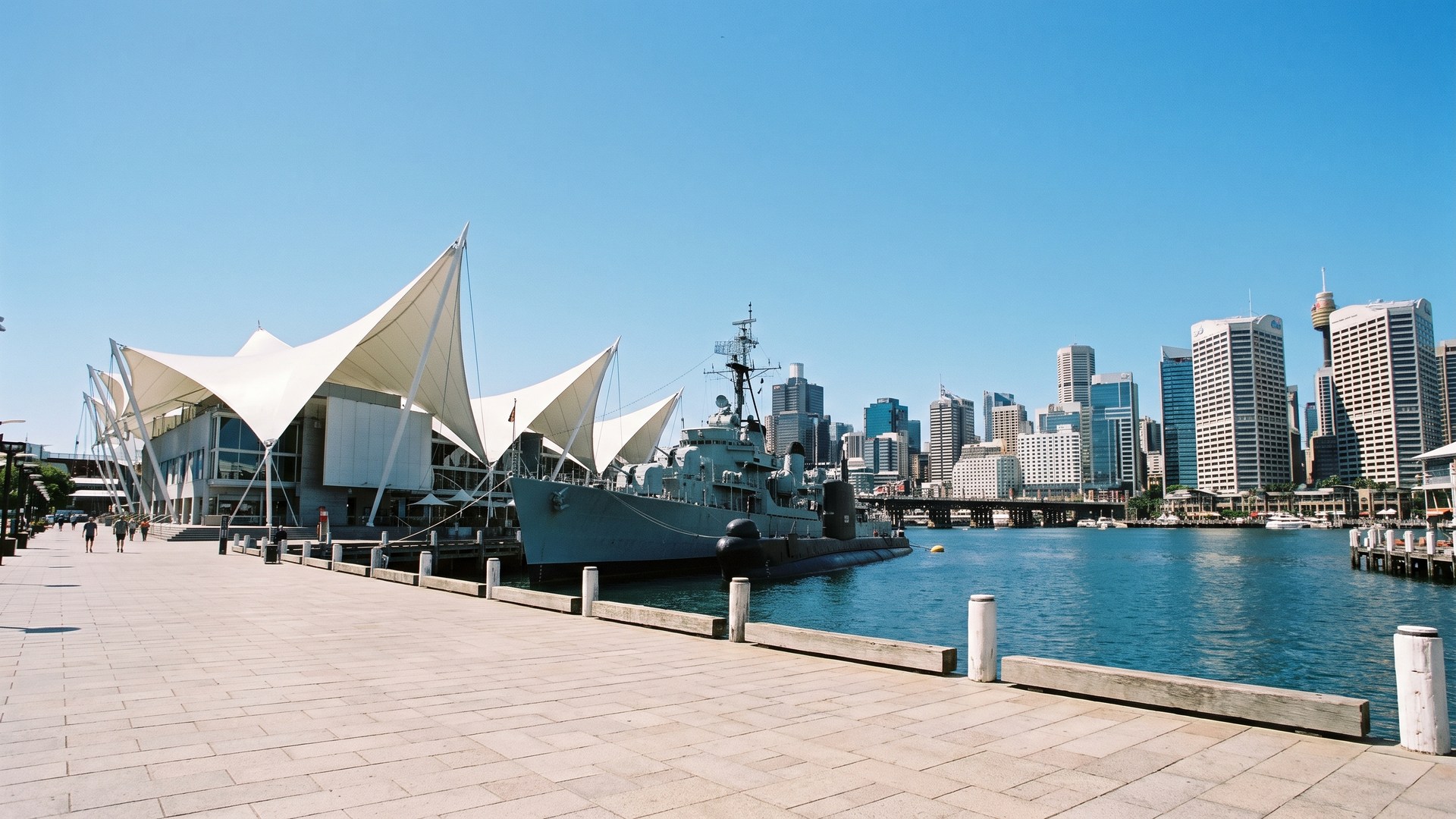 The Darling Harbour waterfront in Sydney, a maritime museum with a white peaked sail roof and a decommissioned naval destroyer moored alongside, the city towers across the water