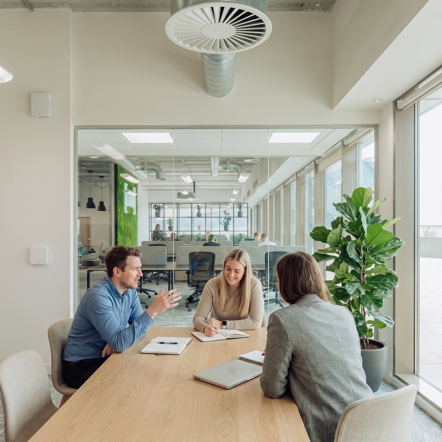 Three colleagues meet at a table in a bright modern office, daylight through glass and a ceiling air diffuser above them.