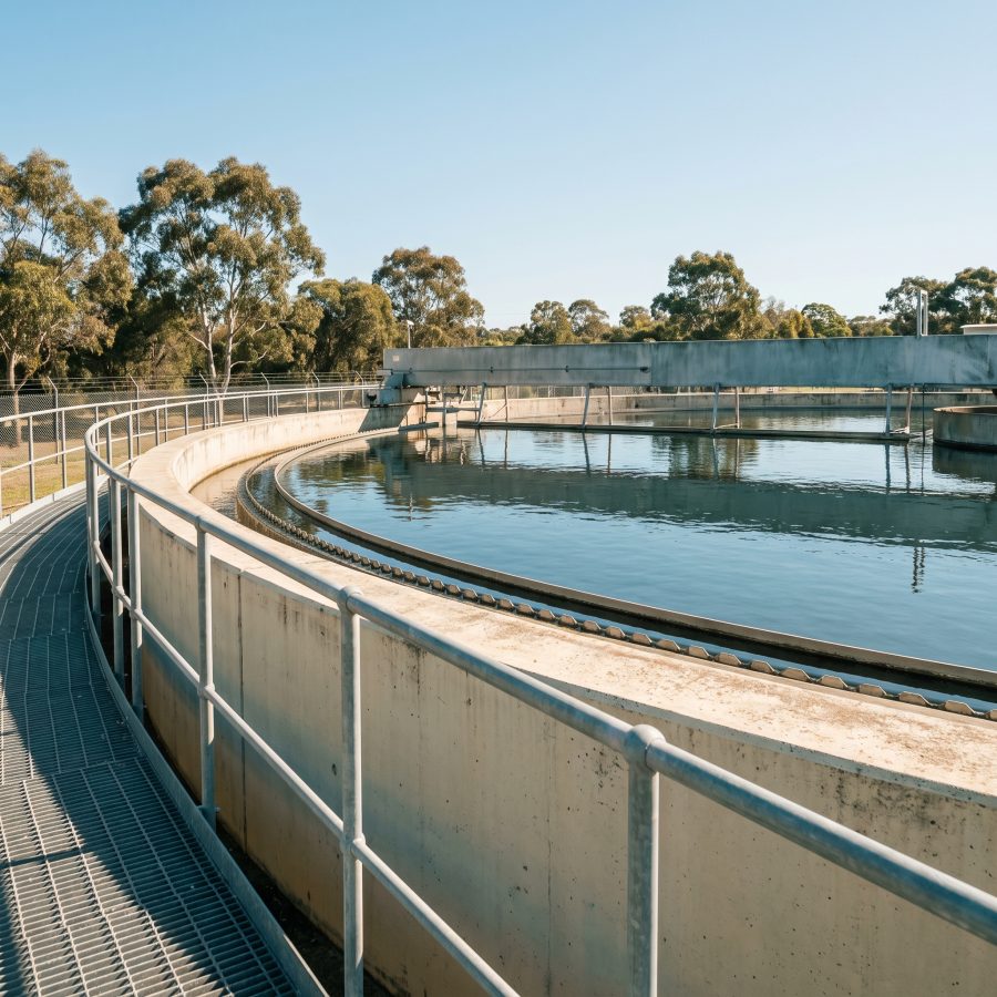 A circular concrete clarifier at a water treatment plant, its handrail walkway curving around the rim under a clear sky.