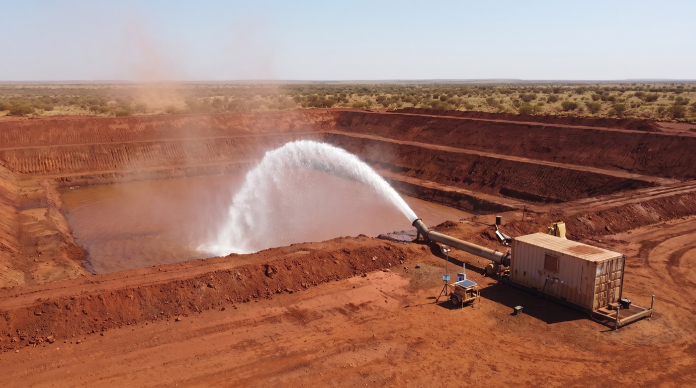 Bore water discharging into a red-earth holding dam at a remote Pilbara mine site, an arid spinifex plain stretching to the horizon