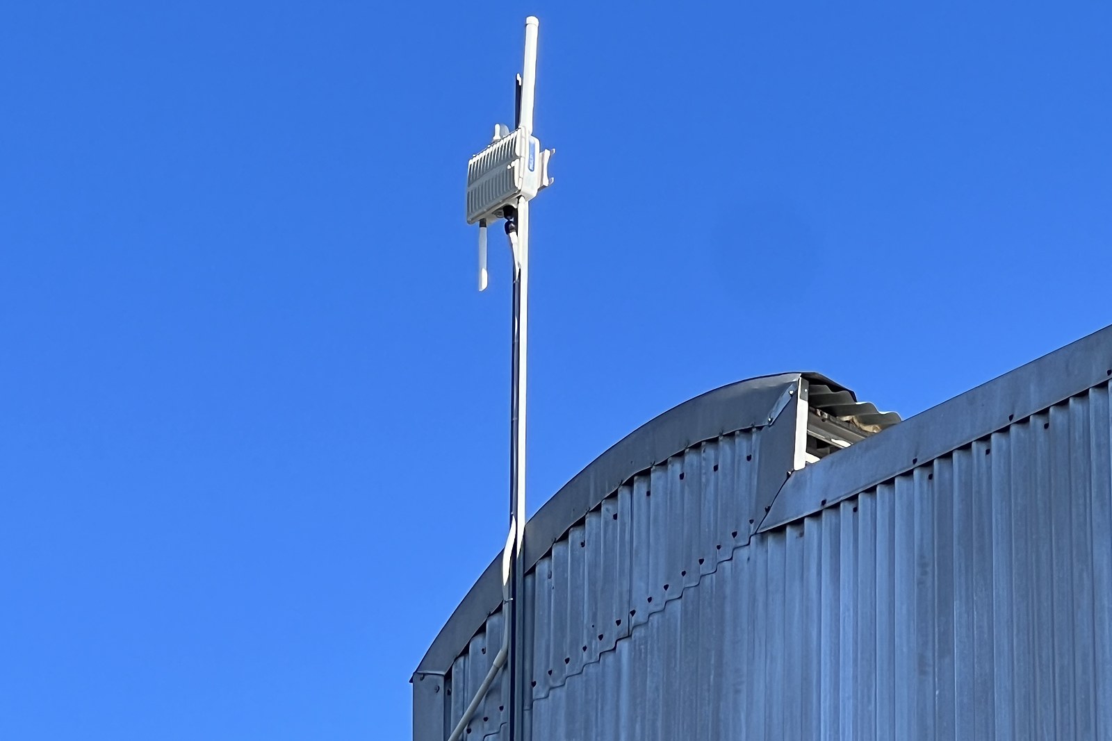 A LoRaWAN gateway enclosure and its antennas mounted on a mast above the curved corrugated roof of a precinct shed