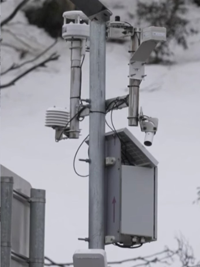 A roadside mast in alpine snow carrying weather sensors, a solar panel and a weatherproof enclosure