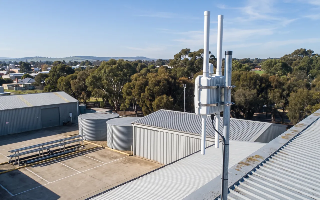 A white outdoor network gateway with two antennas on a galvanised mast above the sheds and water tanks of a works depot, with a town and hills beyond.